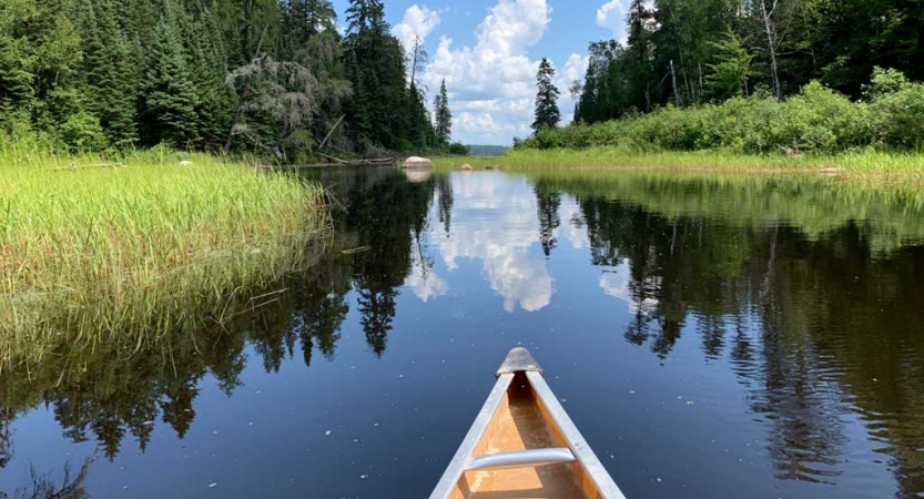 The tip of a canoe appears in the foreground, resting on calm still water that reflects the blue sky dotted with clouds. In the distances, there is a mountainous landscape.